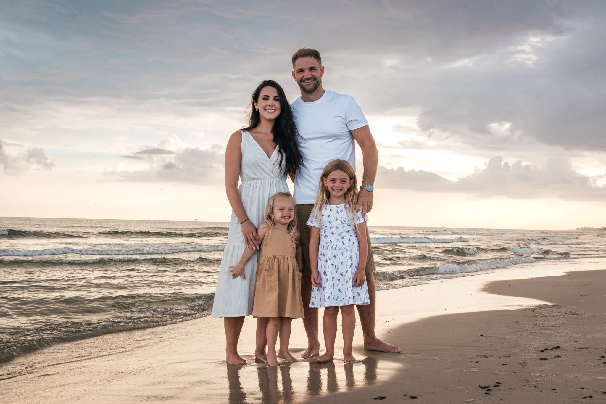 A family of five standing on a beach at sunset, smiling at the camera. The family consists of two adults and three young girls, all dressed in light-colored clothes. The background features ocean waves and a cloudy sky with hints of sunlight.