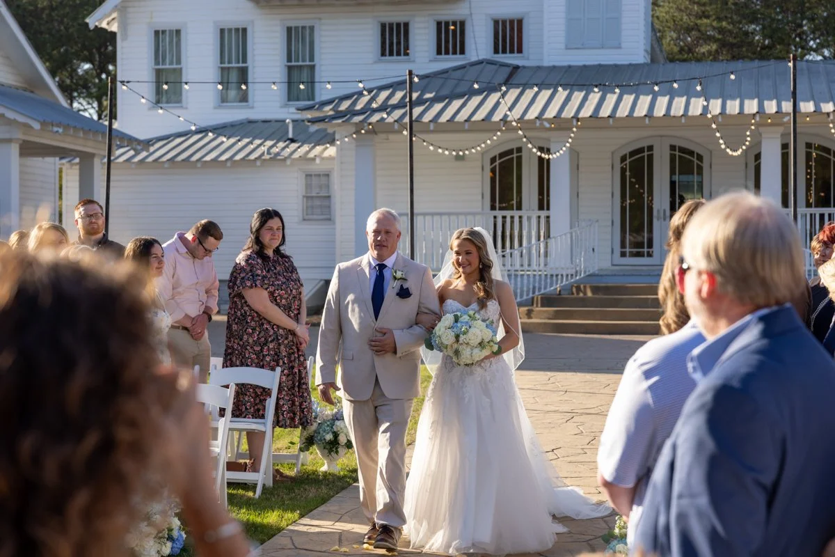 A bride walking down the aisle accompanied by a man, likely her father, during an outdoor wedding ceremony at a white house decorated with string lights.