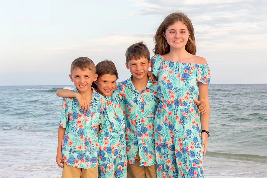 Four children in matching Hawaiian shirts and shorts standing on the beach with ocean waves behind them, smiling and posing.