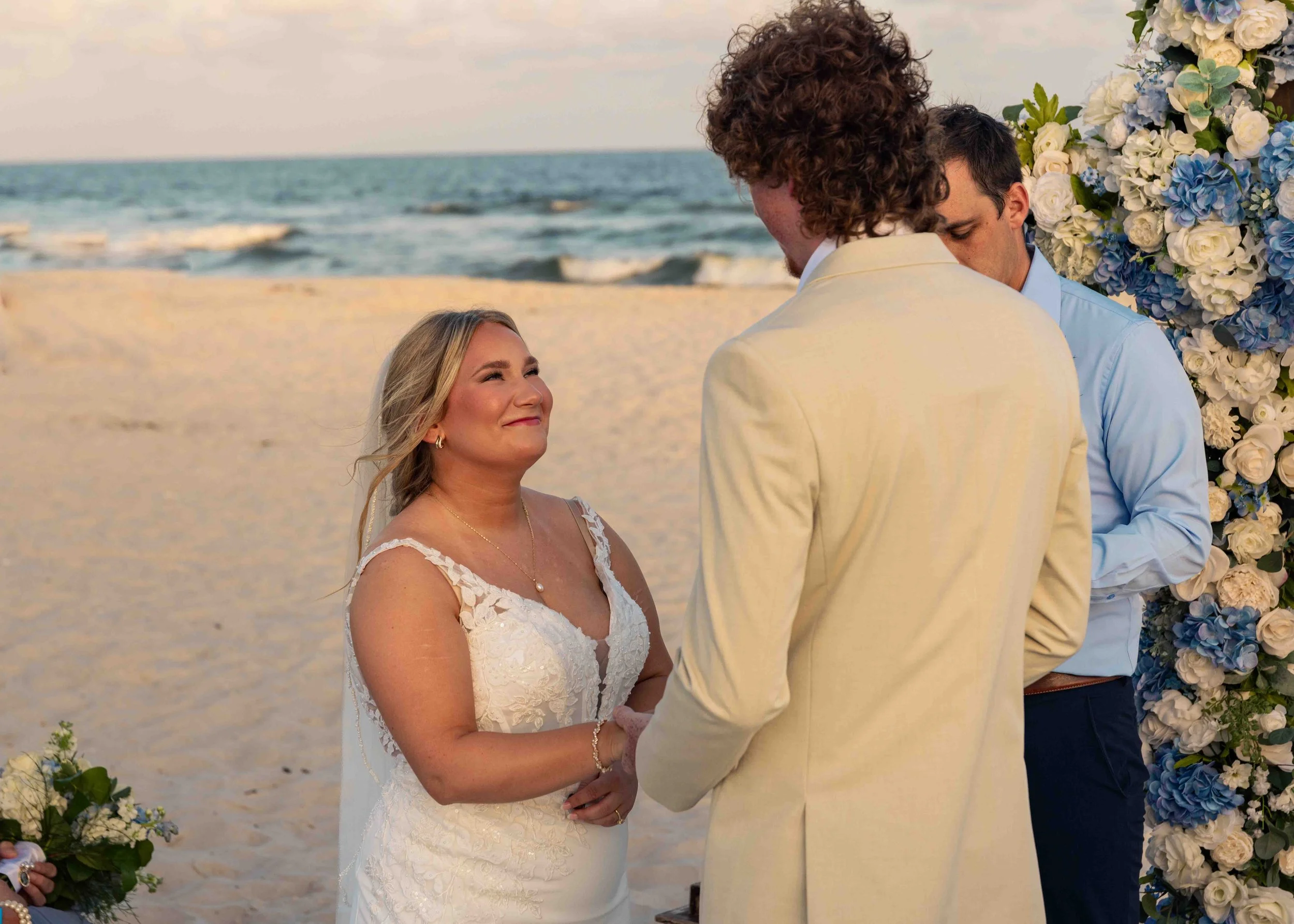 A bride and groom exchange vows during a beach wedding ceremony at sunset, with a floral arch and ocean in the background.