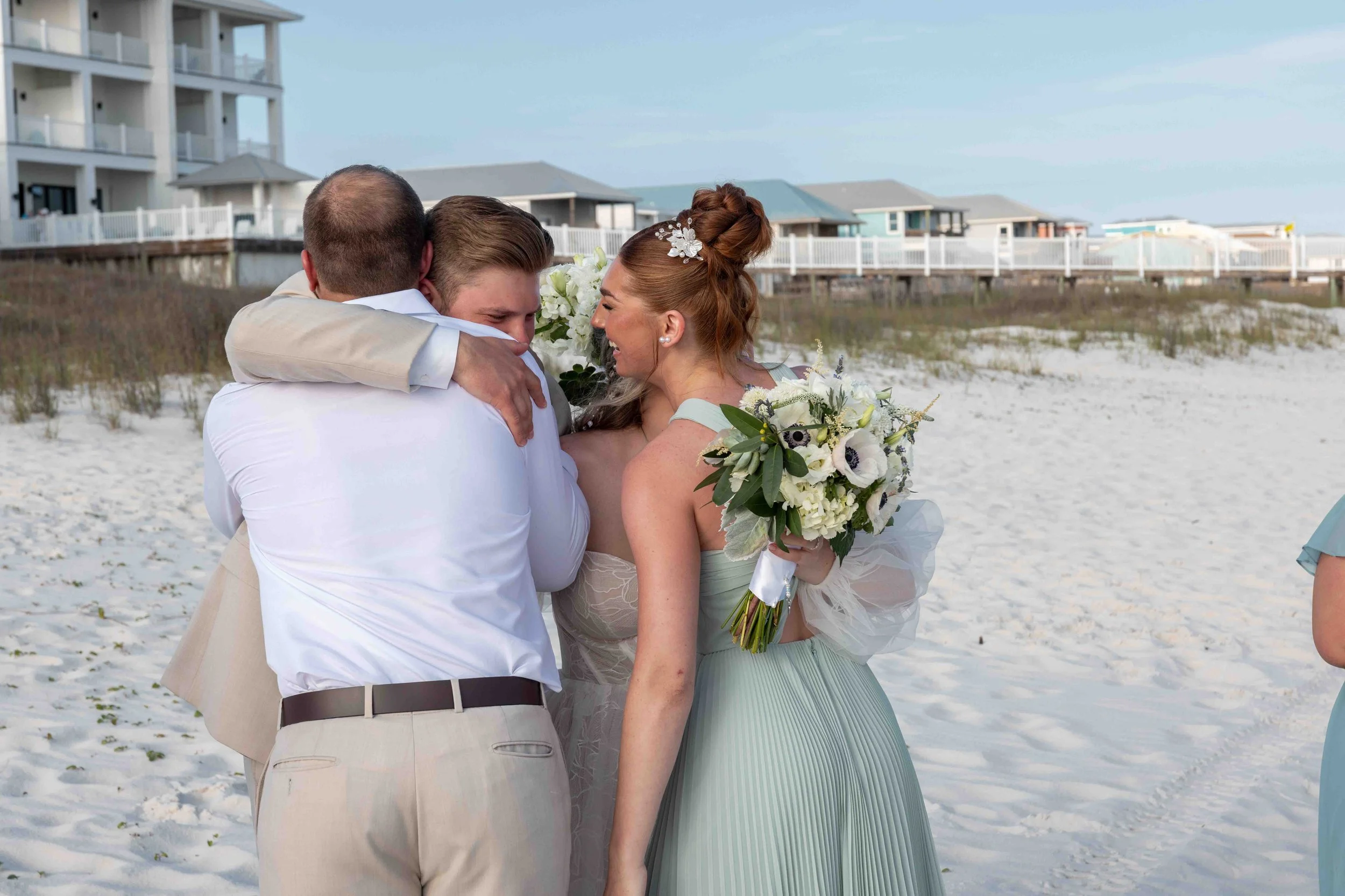 A bride and groom embrace each other outdoors on a beach with beach houses in the background.