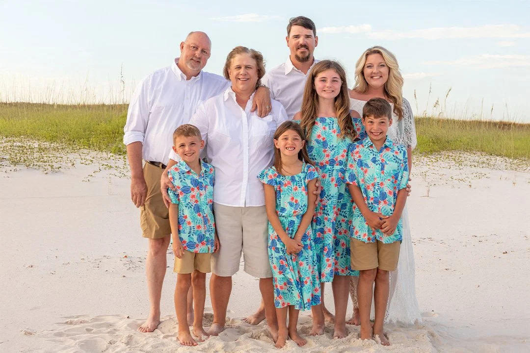 Family of eight on the beach posing for a photo, wearing matching blue floral outfits with sand and beach grass in the background.