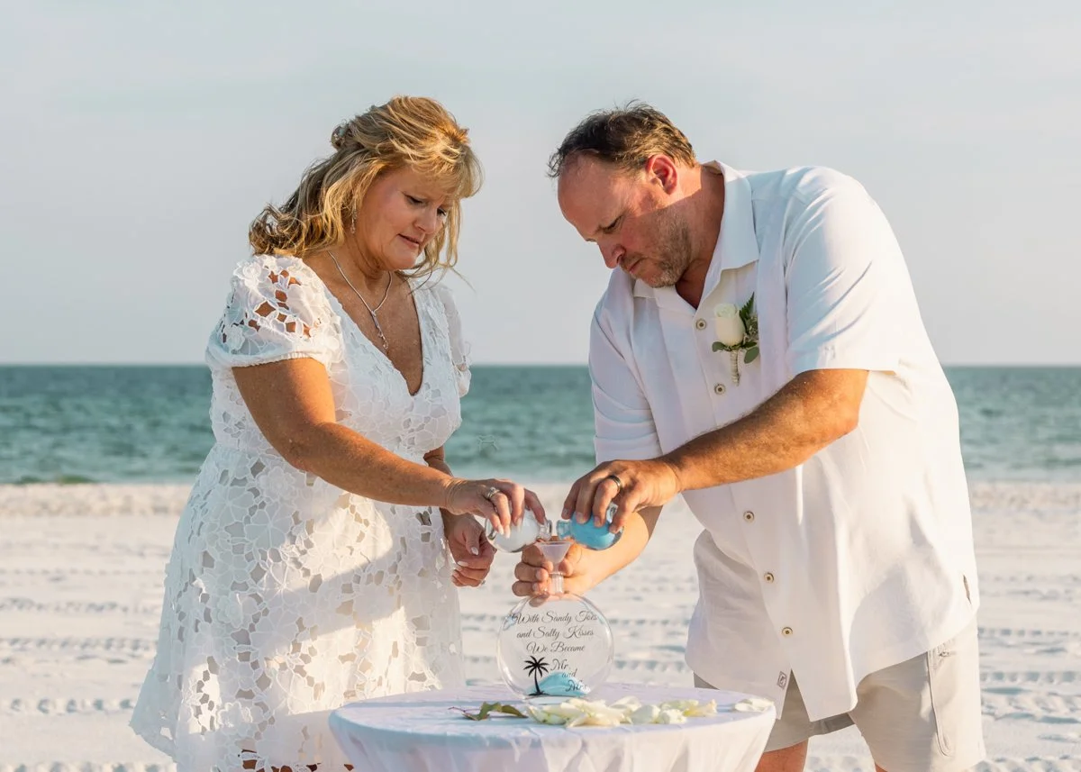 A couple officiating a beach wedding, pouring sand into a glass vase on a table with white flowers, with the ocean in the background.