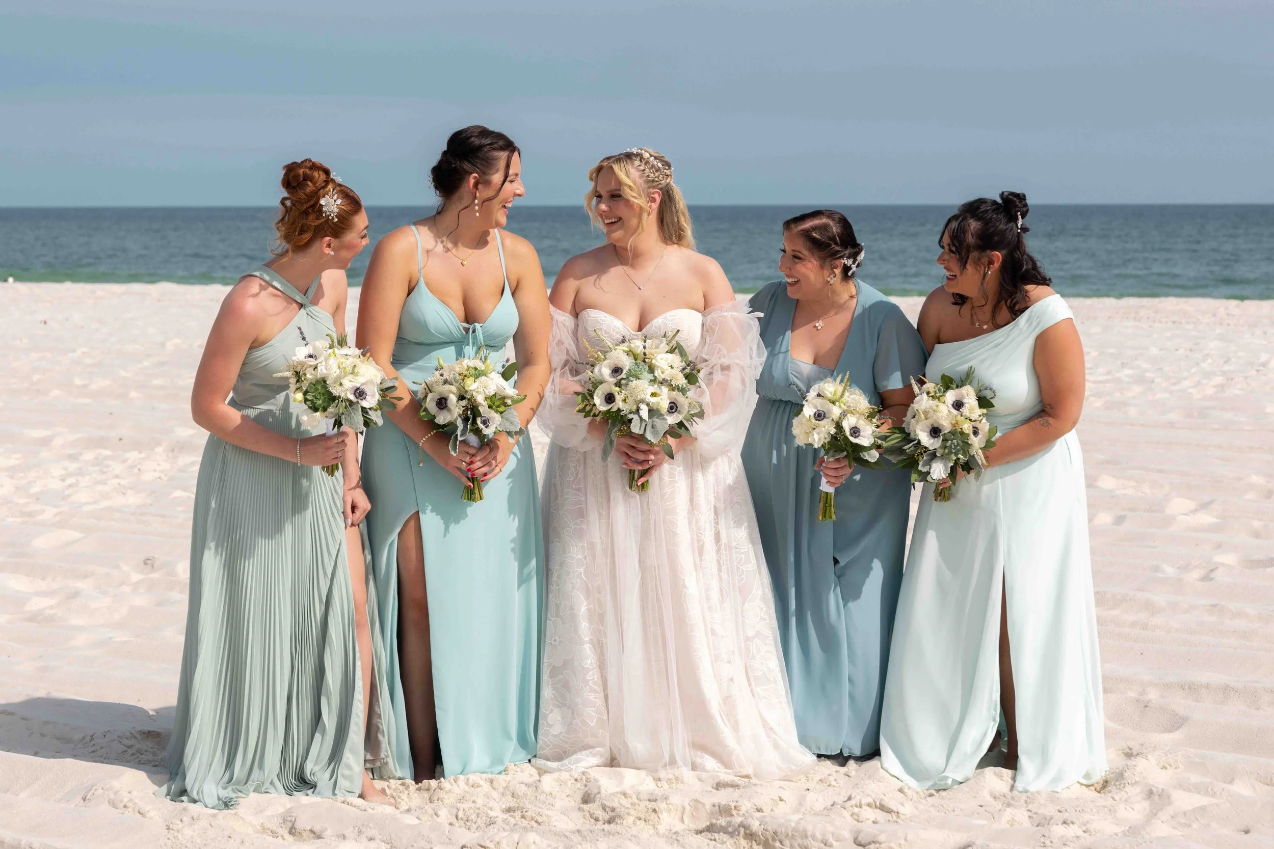 Group of five women dressed in pastel-colored dresses, standing on a sandy beach with the ocean and sky in the background, smiling and holding bouquets of white flowers.