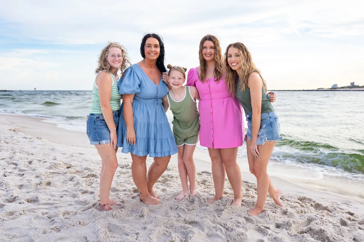 Group of five women standing on a sandy beach near the ocean, smiling at the camera.