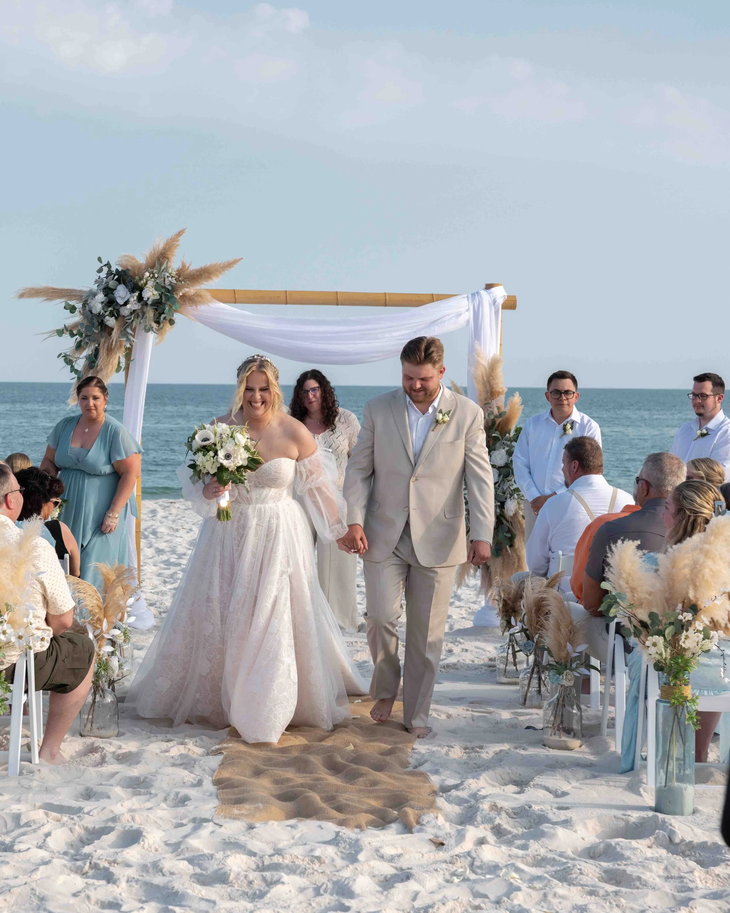 A wedding ceremony taking place on a beach with the ocean in the background, featuring a bride and groom walking hand-in-hand under a decorated arch, surrounded by seated guests.