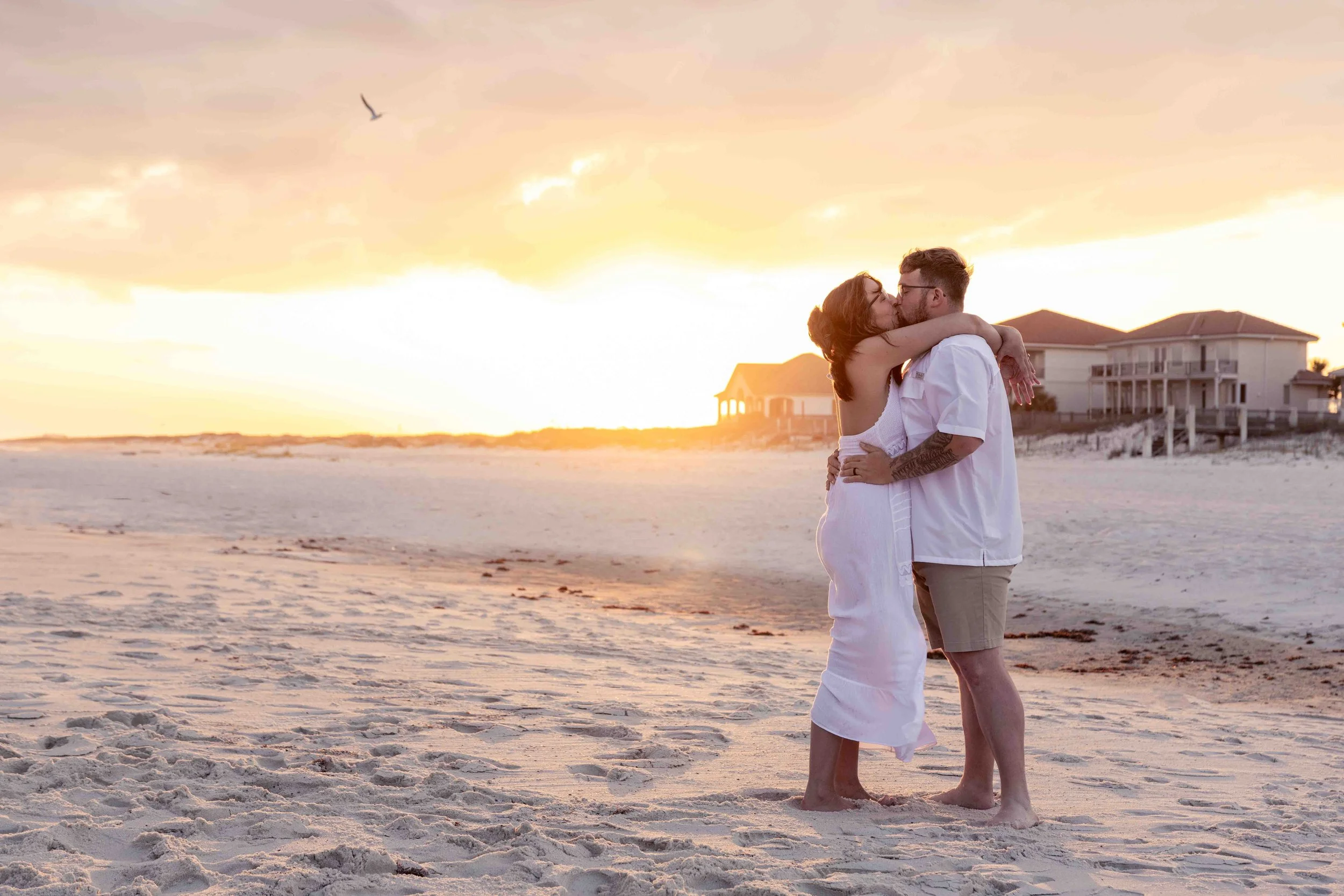 A couple embracing on a beach at sunset, kissing, with houses on stilts in the background and a bird flying in the sky.
