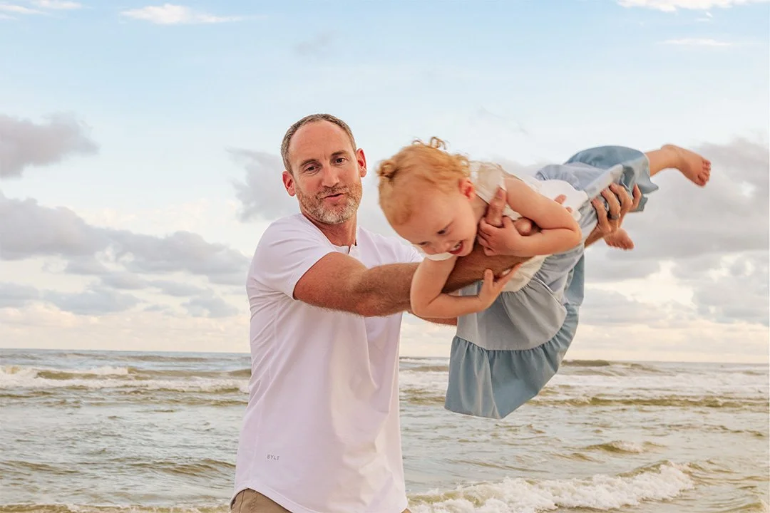 A man holding a young girl in a beach setting with ocean waves and cloudy sky in the background.