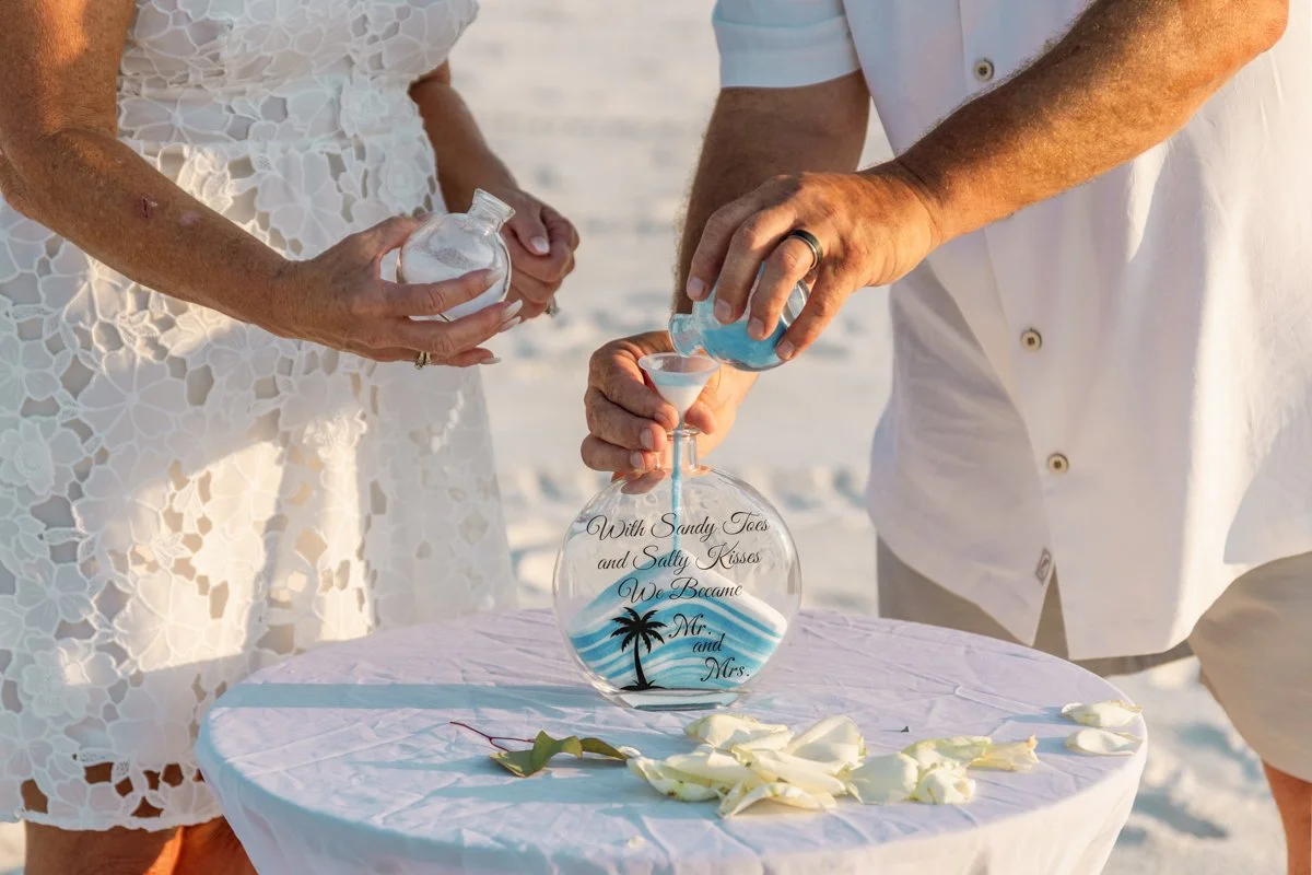 A couple pouring colored sand into a glass bottle during a beach wedding ceremony, with flower petals on a white table.