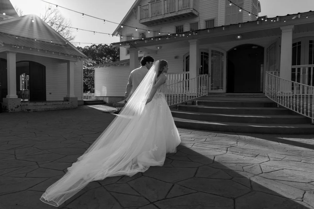 A bride in a wedding gown and veil walking with a groom outside a building with string lights overhead