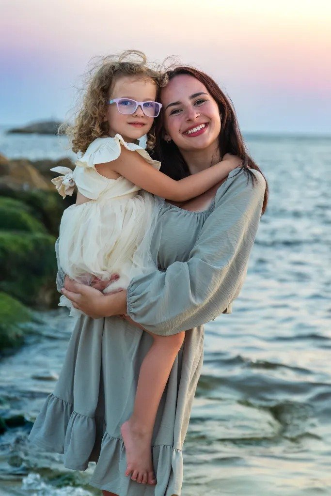 A young woman with long dark hair holding a little girl with curly blonde hair and glasses by the beach during sunset. The woman is smiling, and the girl is hugging her while wearing a cream-colored dress.