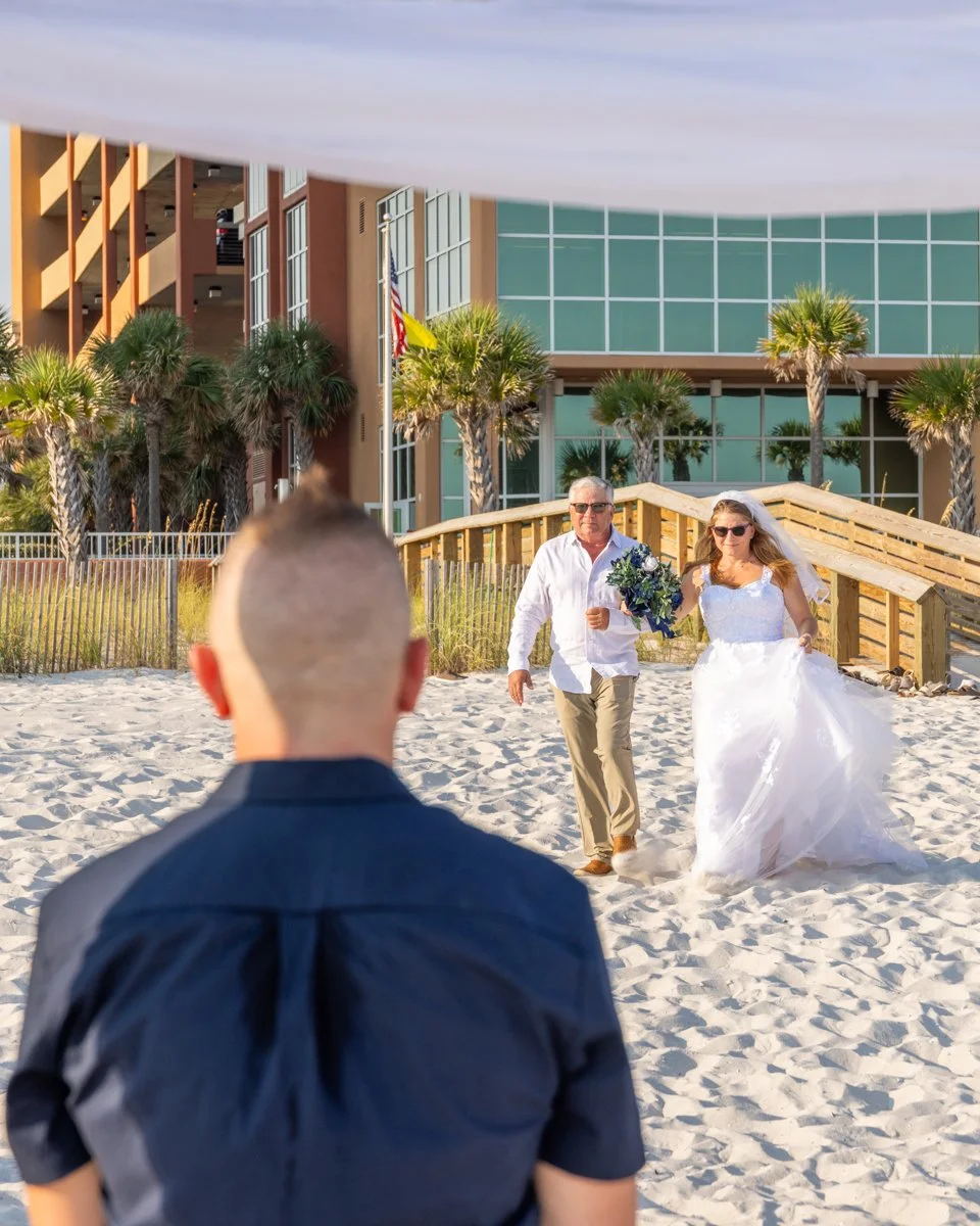 A bride in a white wedding dress walking on the beach with a man carrying a bouquet, approaching a man in a black shirt, with a hotel and palm trees in the background.