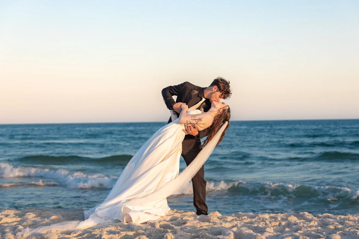 A man and woman in wedding attire sharing a kiss on a beach, with the ocean and a clear sky in the background.