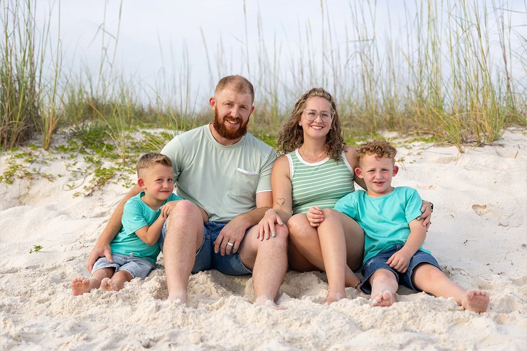 A family of four sitting on a sandy beach with tall grass behind them, smiling at the camera.