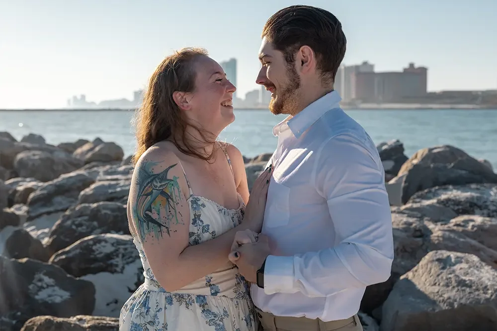 A couple standing close together on rocks near the water, smiling at each other with city buildings in the background.