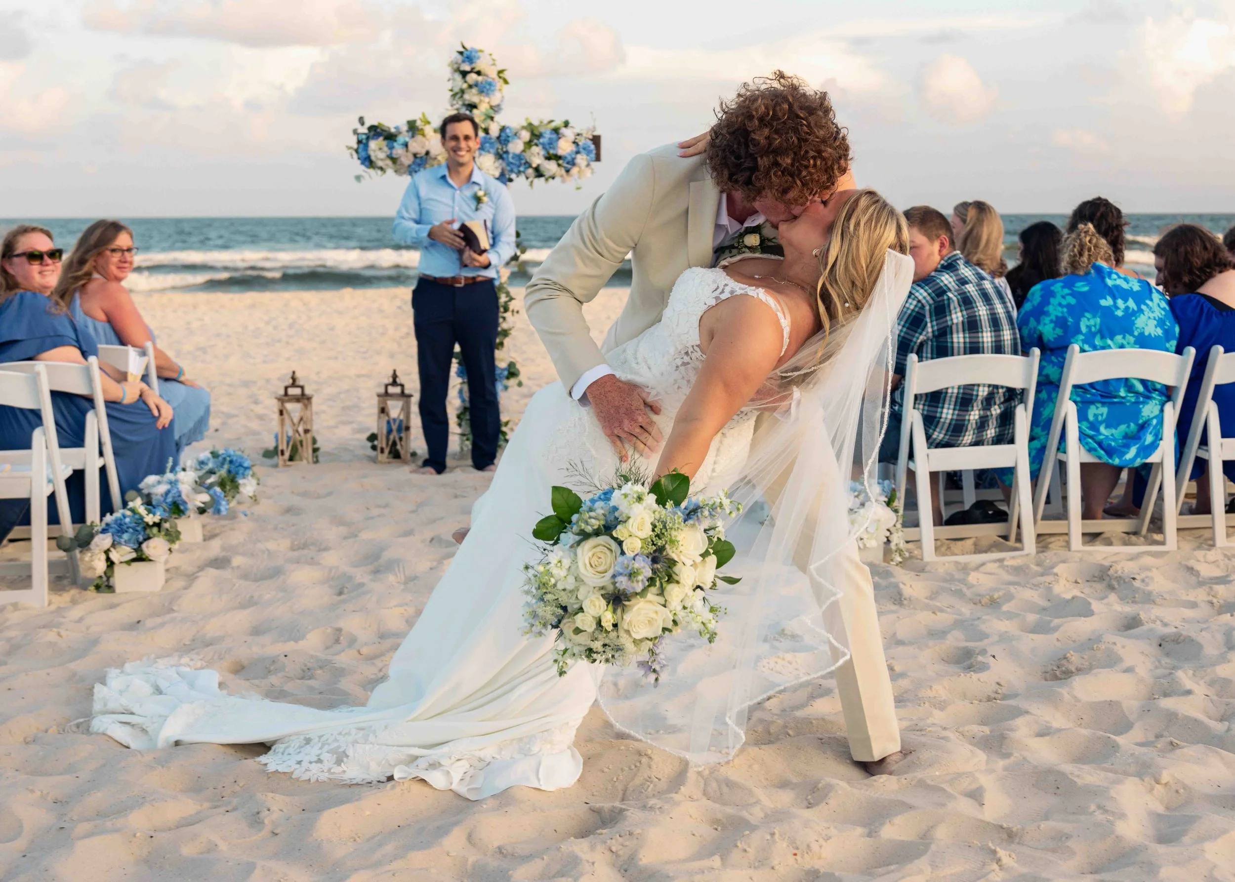 A wedding ceremony on the beach with a couple kissing, surrounded by guests seated on white chairs, decorated with flowers, with the ocean and sky in the background.
