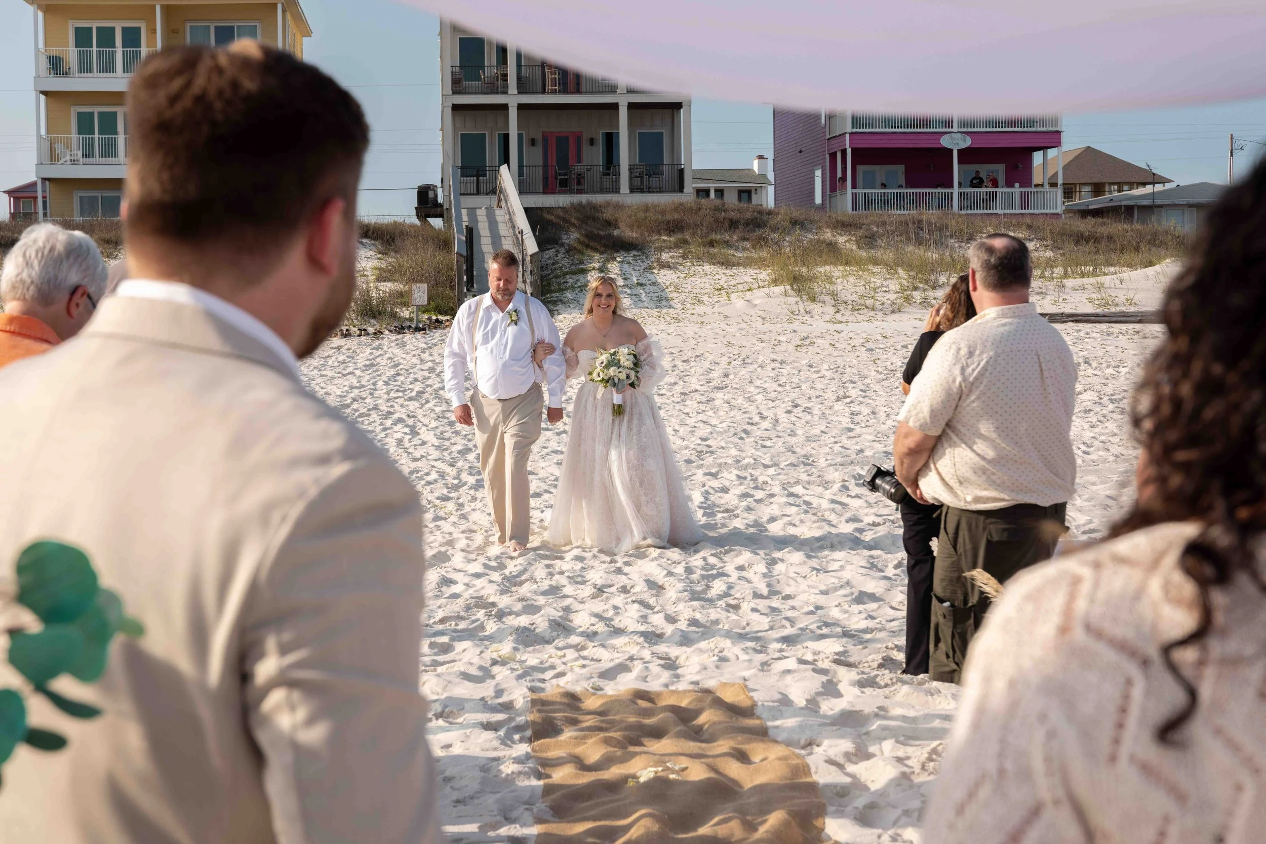 A bride and groom walking on a sandy beach during a wedding ceremony, with guests and beach houses in the background.