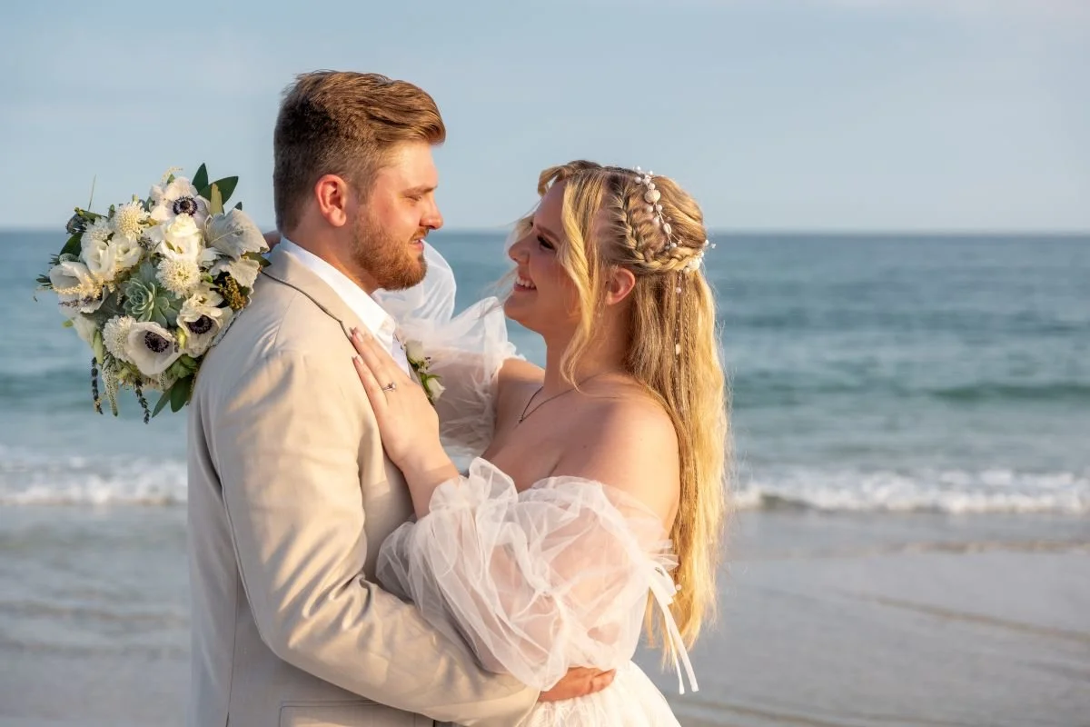 A bride and groom standing close together on a beach, smiling at each other, with the ocean in the background. The groom is holding a bouquet of white flowers.