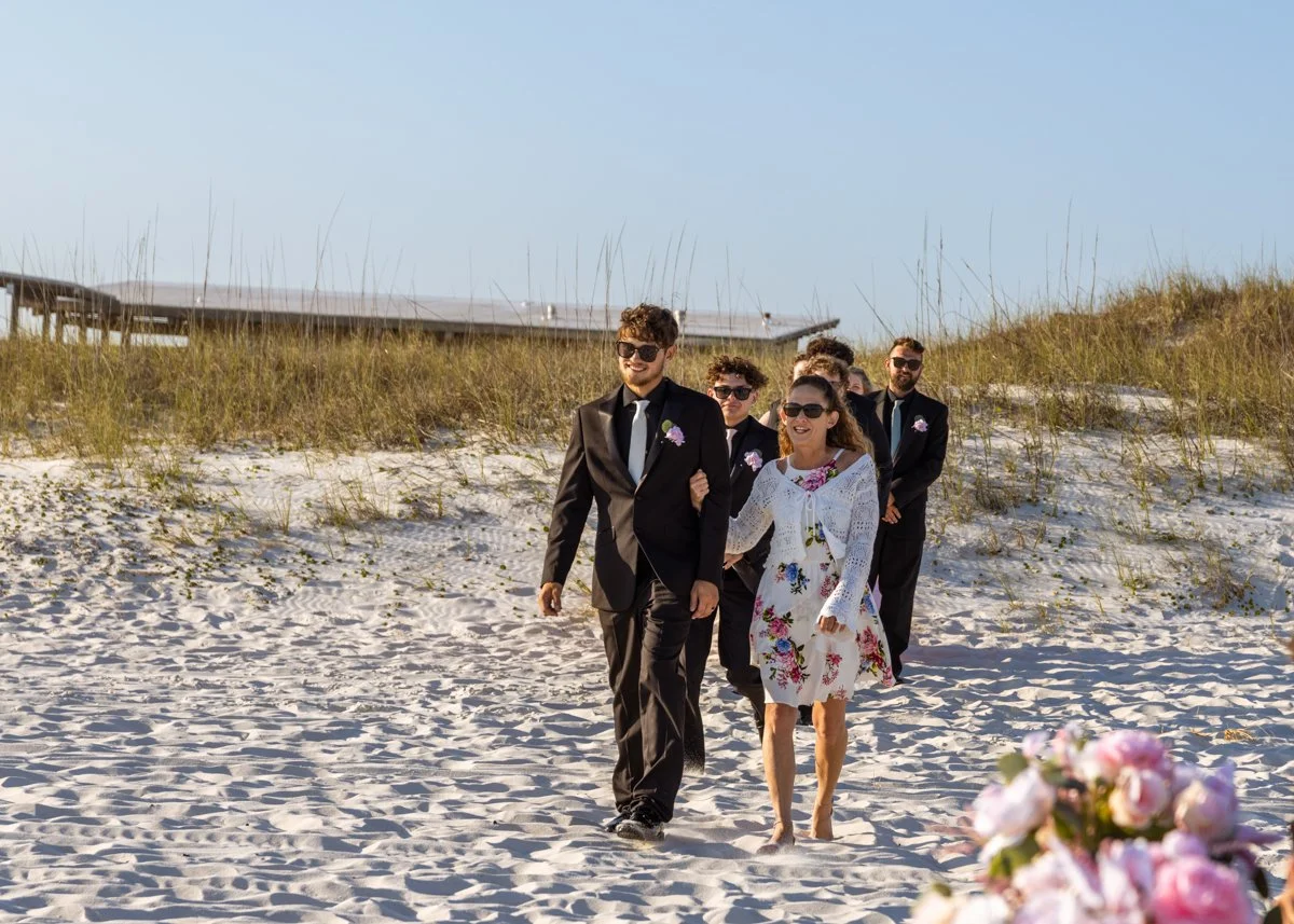 Group of six people walking on the beach, dressed in formal attire, with some wearing sunglasses and smiling, and a blurred bouquet of pink flowers in the foreground.