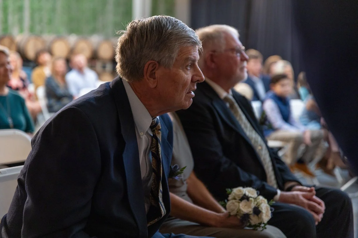 Two elderly men in suits sitting side by side in a formal event, holding bouquets of white flowers, with a blurred audience in the background.
