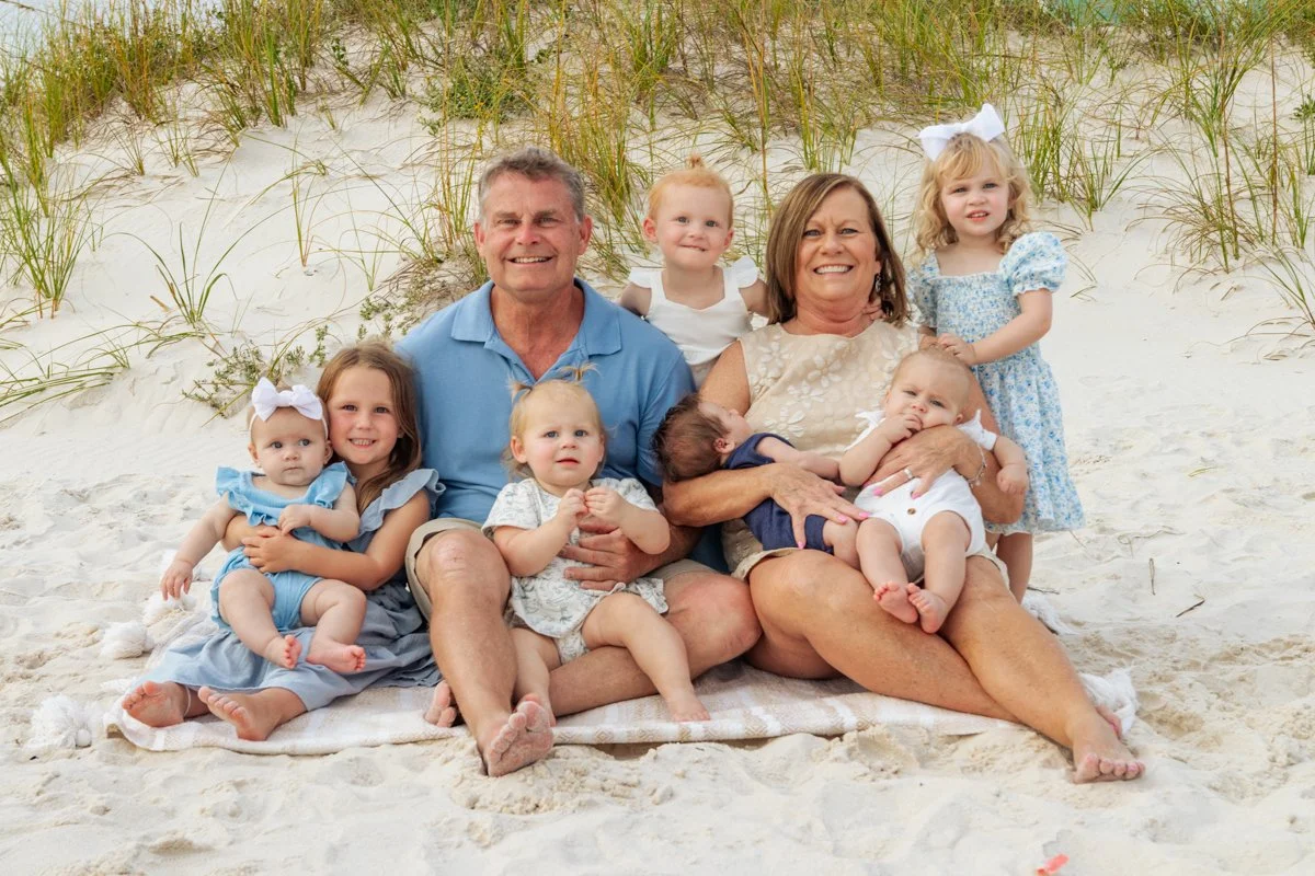 A family with seven children sitting on the sand at a beach with grassy dunes in the background, smiling for a photo.