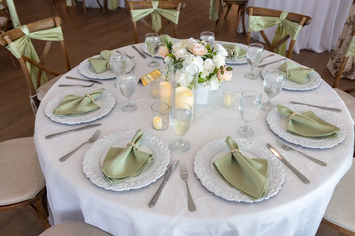 Round banquet table set for a formal event with white tablecloth, white decorative plates, light green napkins with pearl napkin rings, wine glasses, and silverware. A floral centerpiece with white and blush flowers, surrounded by candles, decorates 