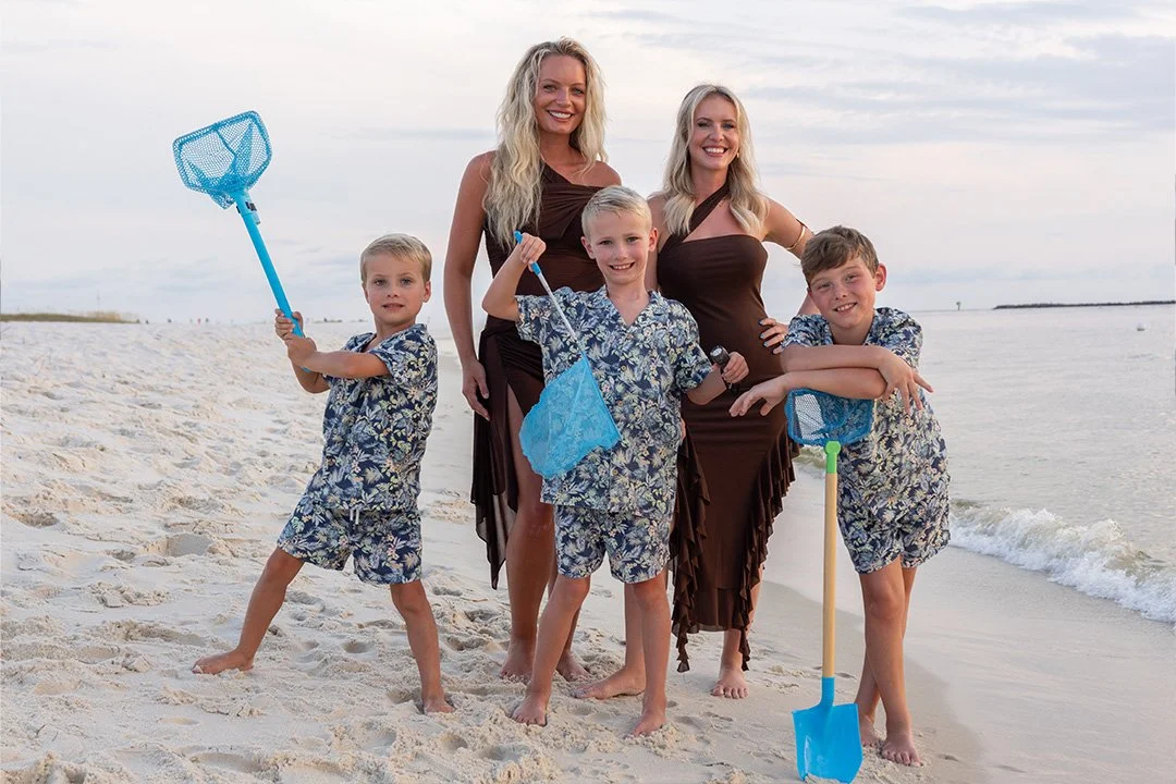 A family of two women and three boys standing on a sandy beach with a calm ocean in the background. They are smiling and holding beach toys, including a net, a butterfly net, and sand shovels, dressed in matching floral swimwear and brown dresses.