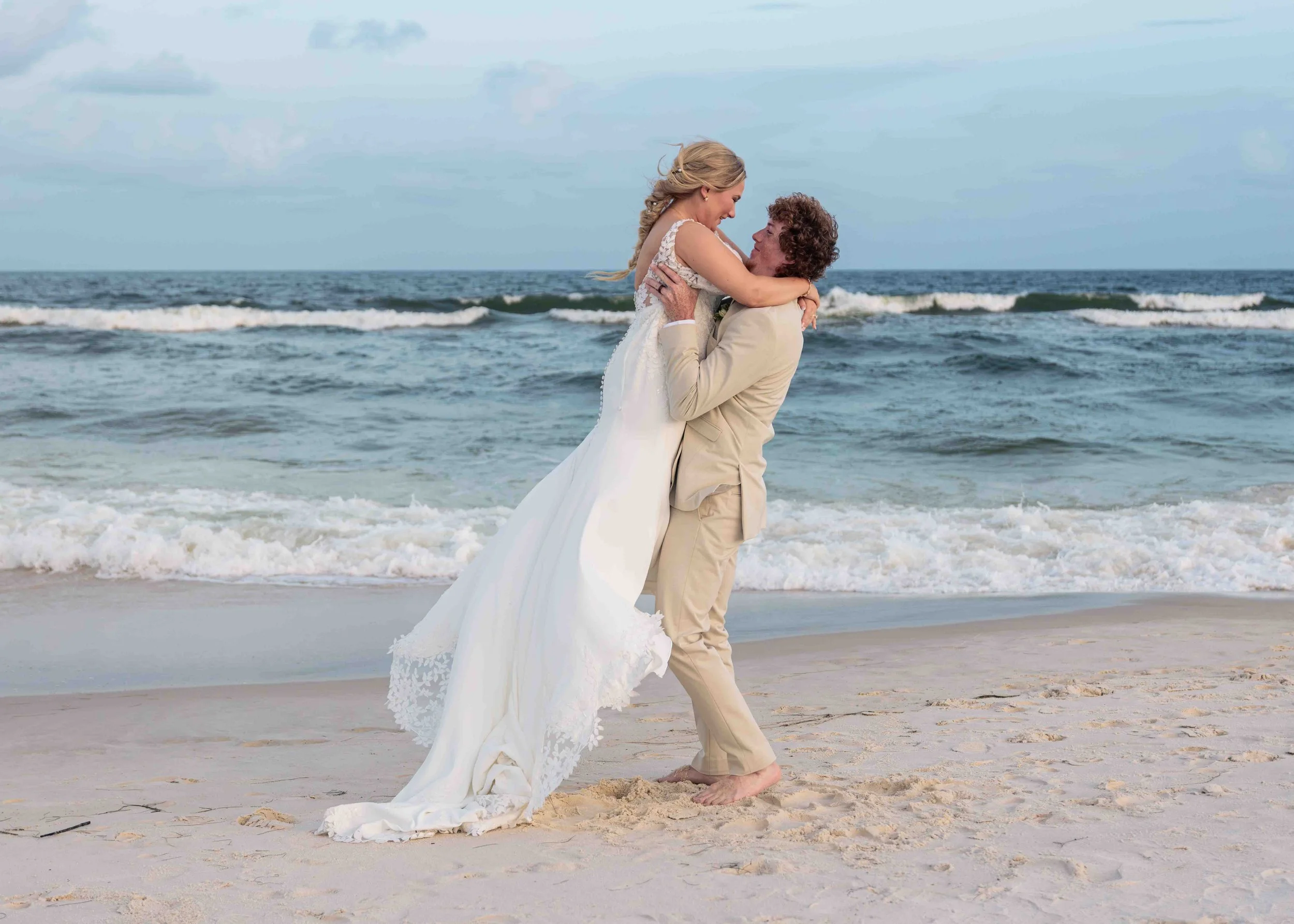A bride and groom on the beach, with the groom lifting the bride in his arms. The woman wears a white wedding dress and the man wears a tan suit. They are smiling and engaging in a romantic moment near the ocean.