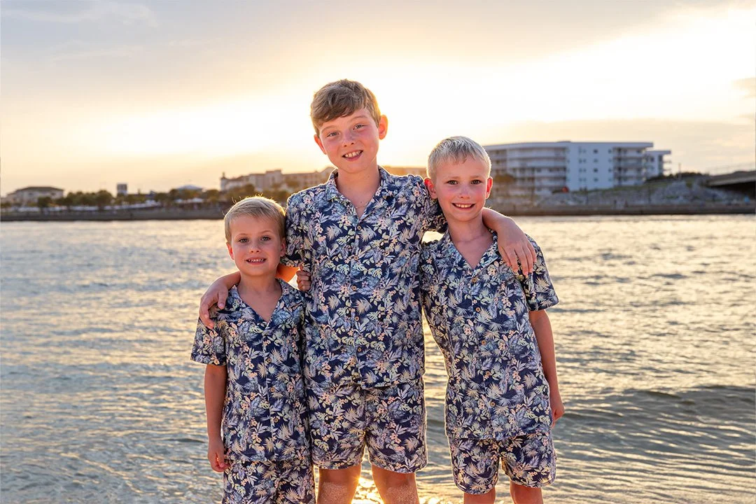 Three boys in matching floral shirts and shorts standing by the water at sunset, with buildings in the distance.