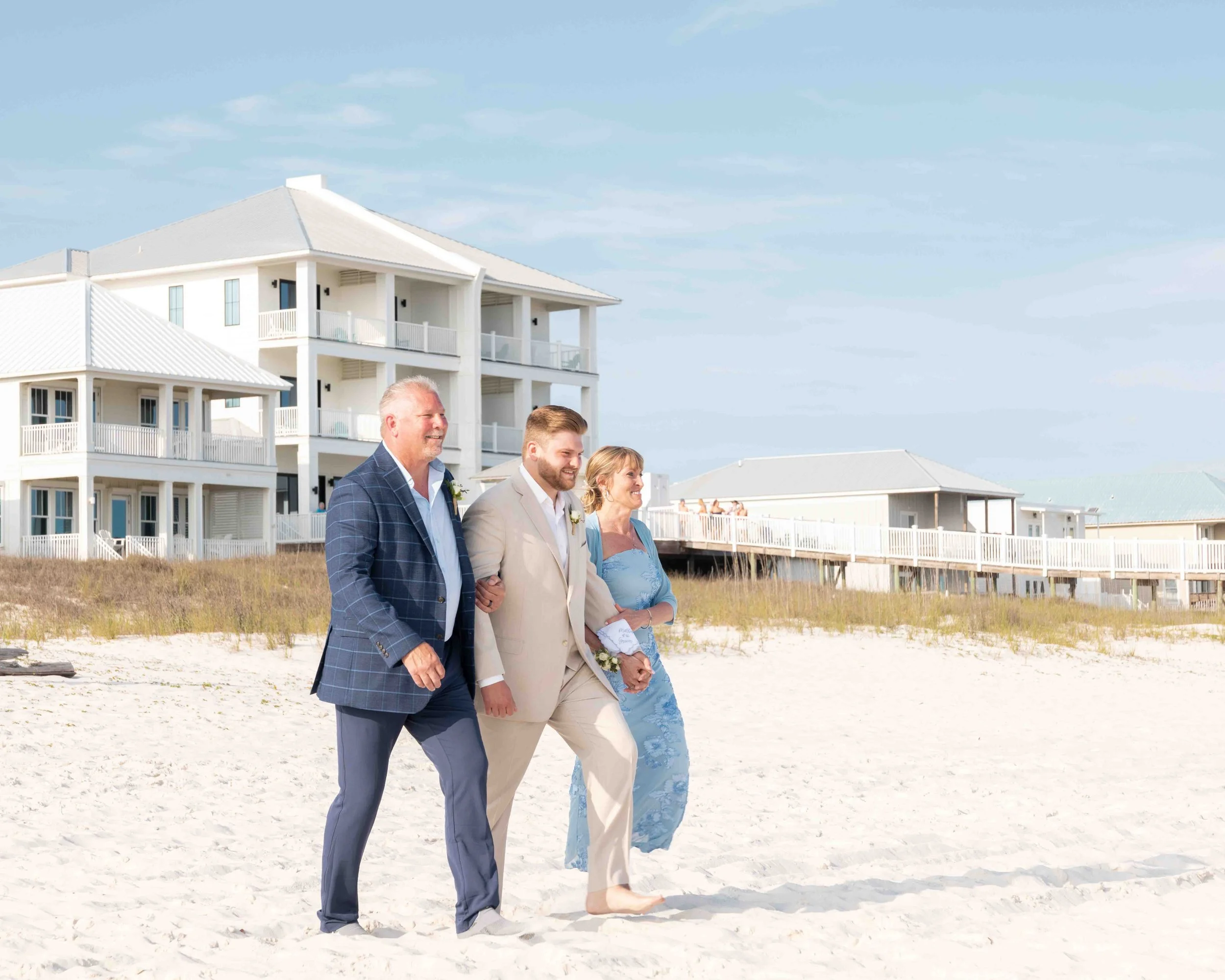 Four people dressed in formal attire walk along a sandy beach with white, multi-story houses on stilts behind them, under a clear blue sky.