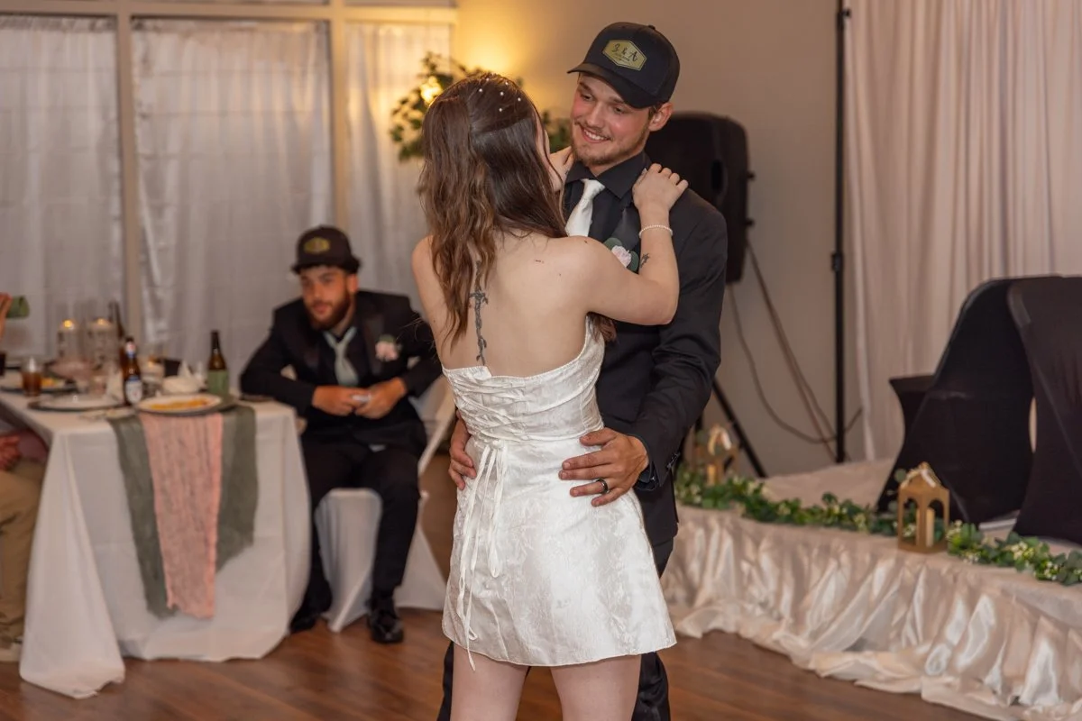 Couple dancing at a wedding reception, the woman in a satin white dress and the man in a suit with a black hat, smiling and holding each other.