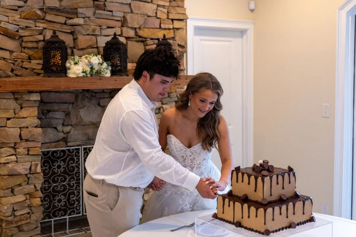 A bride and groom cutting a two-tier wedding cake with chocolate drizzle in a cozy room with a stone fireplace and decorative lanterns.