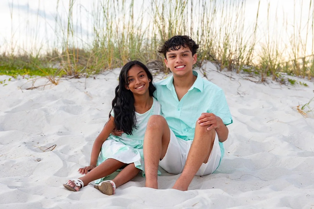 A boy and a girl sitting on the sand at the beach, smiling at the camera with tall grass behind them.