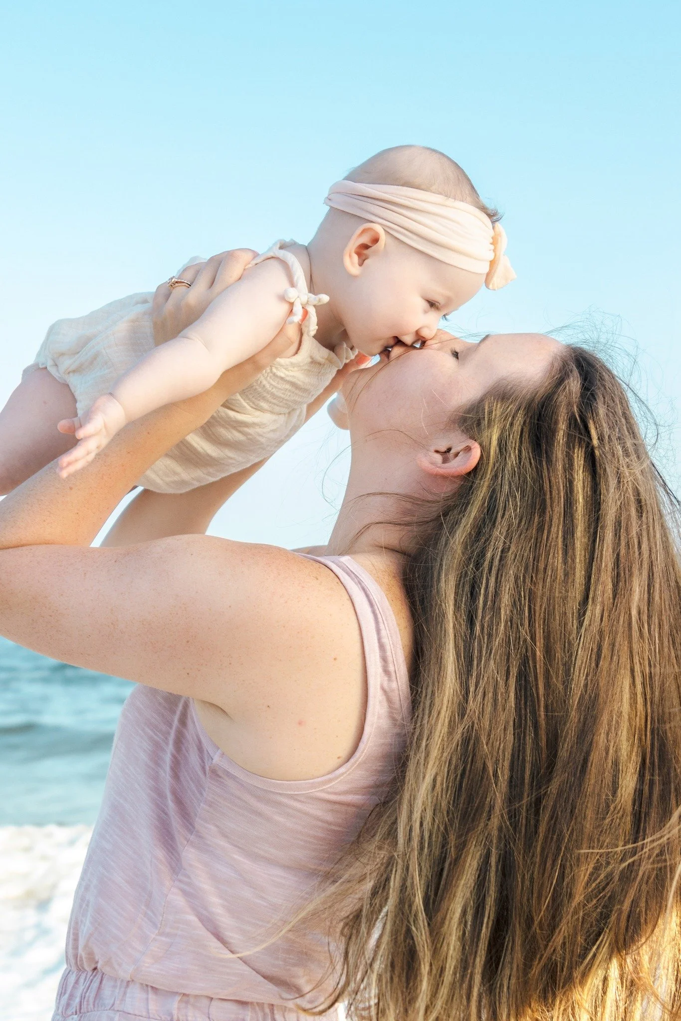 A woman with long hair holding a baby girl at the beach. The baby is wearing a headband and a light-colored outfit. They are sharing a kiss with the ocean in the background.