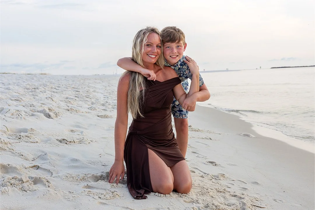 A woman kneeling on the beach with a young boy on her back, both smiling.
