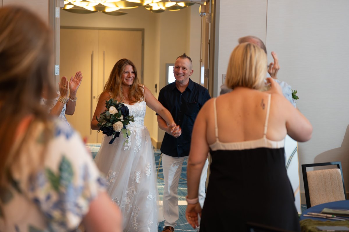 A bride in a white wedding dress holding a bouquet and smiling, walking into a room with friends and family greeting her, some clapping and waving.