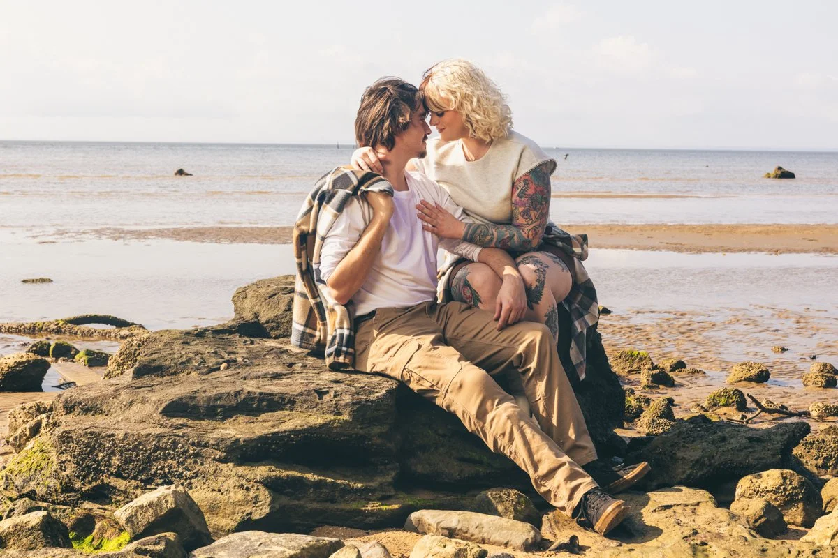 A couple sitting closely on rocks by the beach, gazing into each other's eyes with the ocean in the background.
