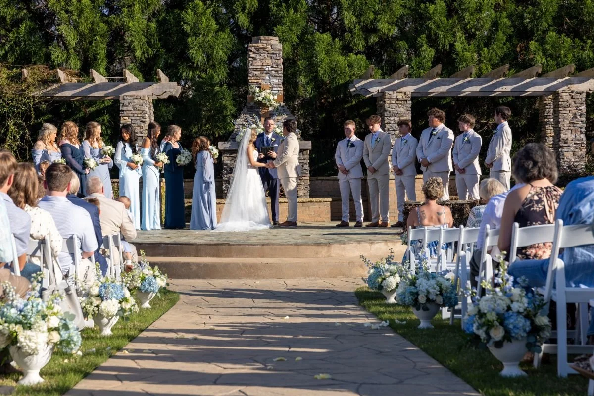 A wedding ceremony taking place outdoors on a wooden platform with a bride in a white gown and veil, and a groom in a light-colored suit, exchanging vows. Bridesmaids in light blue and dark blue dresses stand on the left, and groomsmen in gray suits 