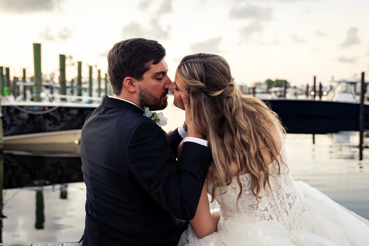 A bride and groom share an intimate moment near a marina, with boats in the background during sunset.