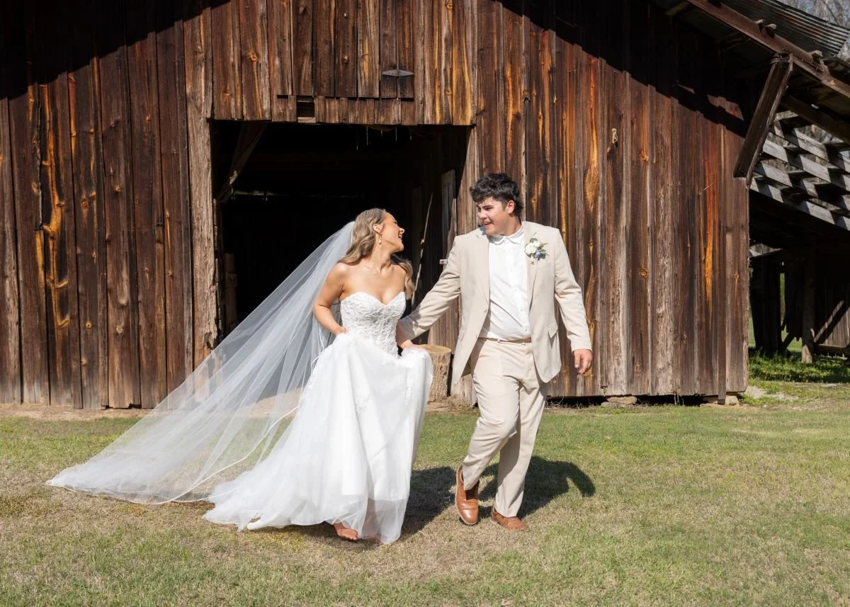 Bride and groom walking hand in hand outside a rustic wooden barn, smiling and looking at each other.