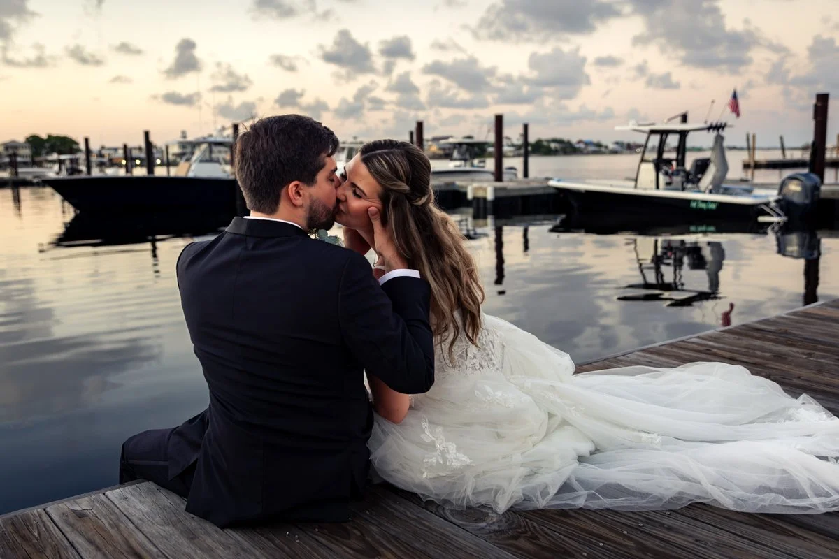 A newlywed couple sharing a kiss on a dock by the water during sunset, with boats in the background.
