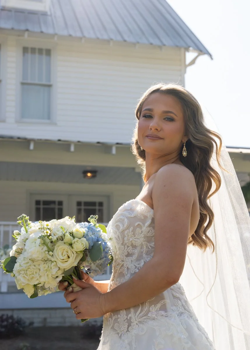 A bride with long wavy hair wearing a white wedding dress with lace details and earrings, holding a bouquet of white and pale blue flowers, standing outdoors in front of a white house with a metal roof.