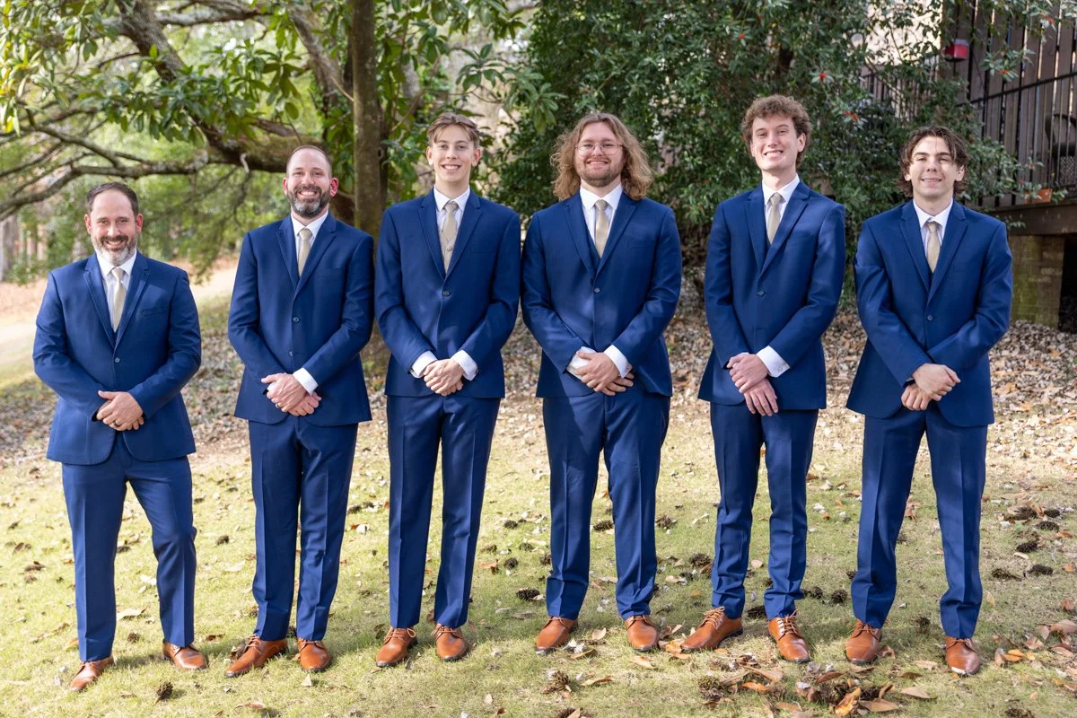 Six men dressed in blue suits and beige ties standing outdoors on grass with trees in the background.