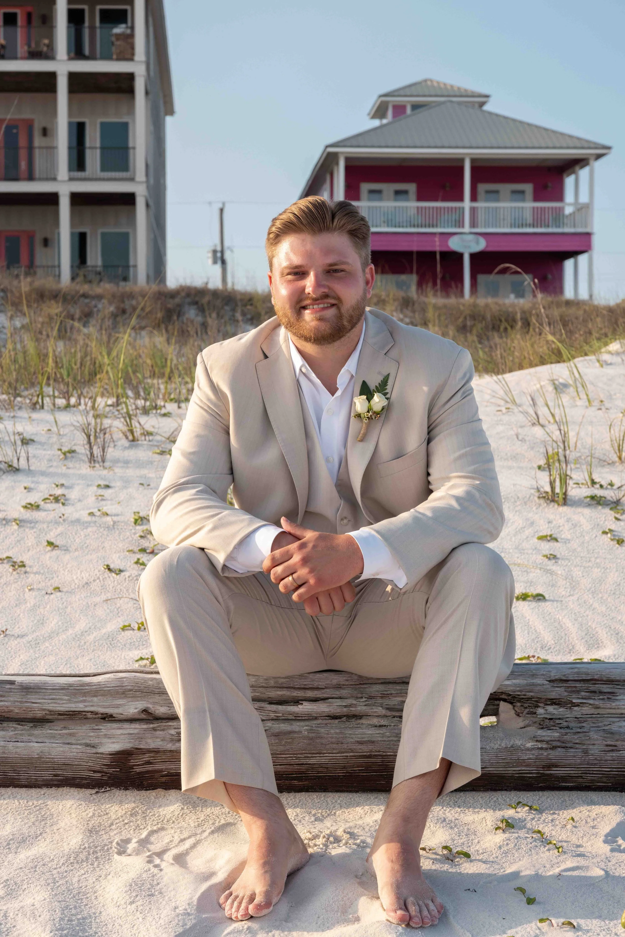 A man in a beige suit with a white shirt and boutonniere sitting on a wooden log on a sandy beach with grass, with colorful houses in the background.