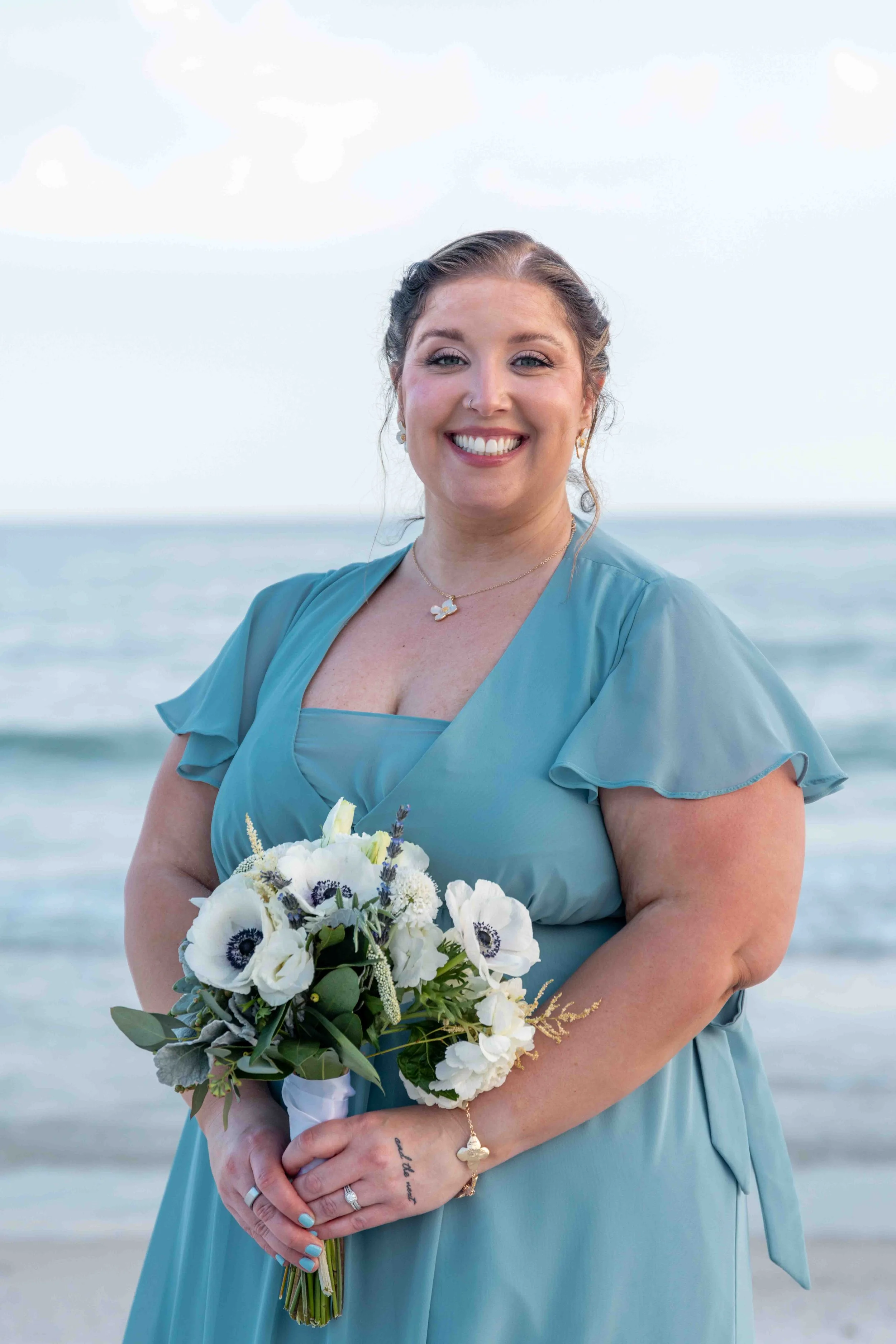 A woman in a blue dress smiling at the camera while holding a bouquet of white flowers with purple accents, standing on the beach near the ocean.