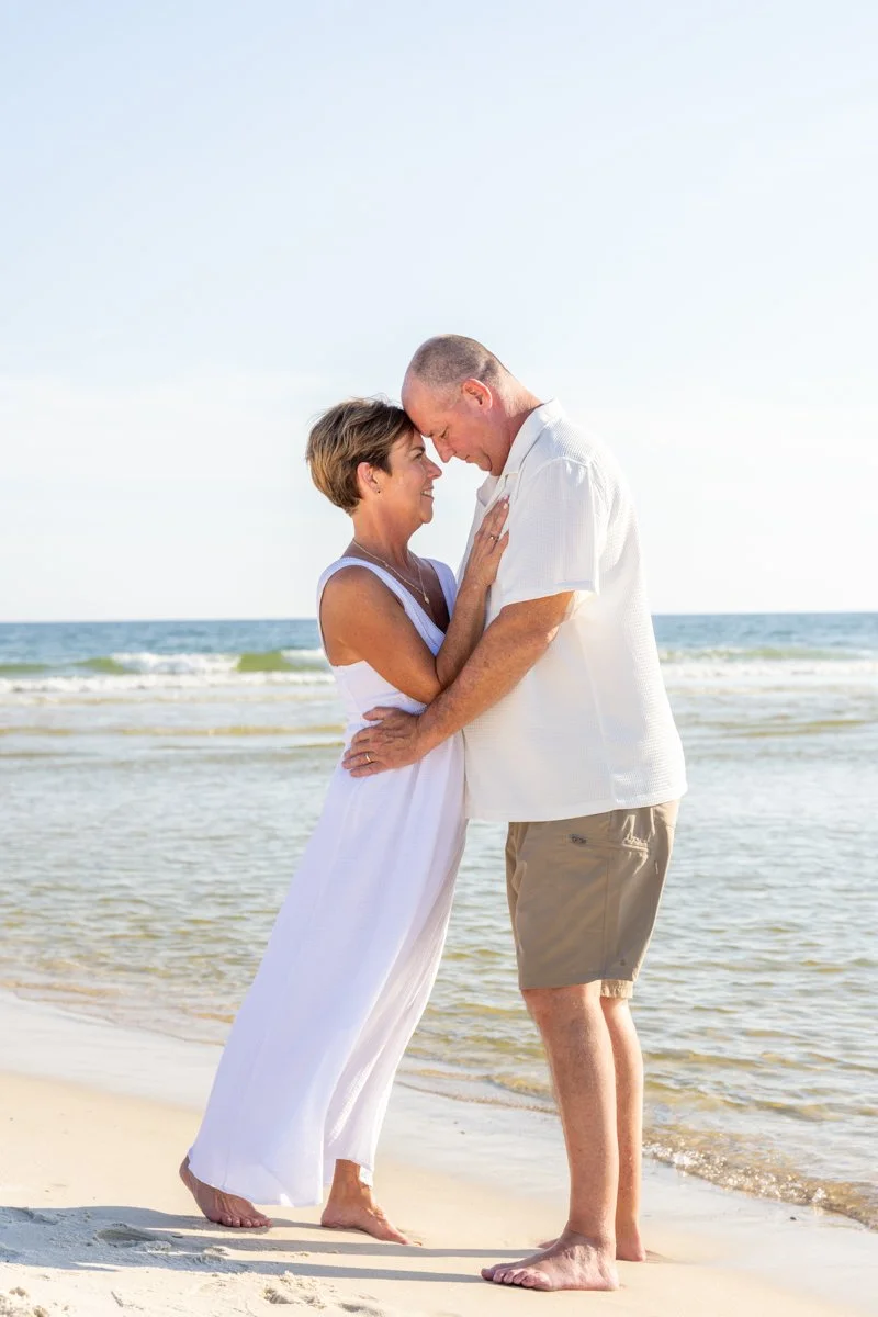 A couple standing close together on the beach, touching foreheads and smiling, with the ocean in the background.