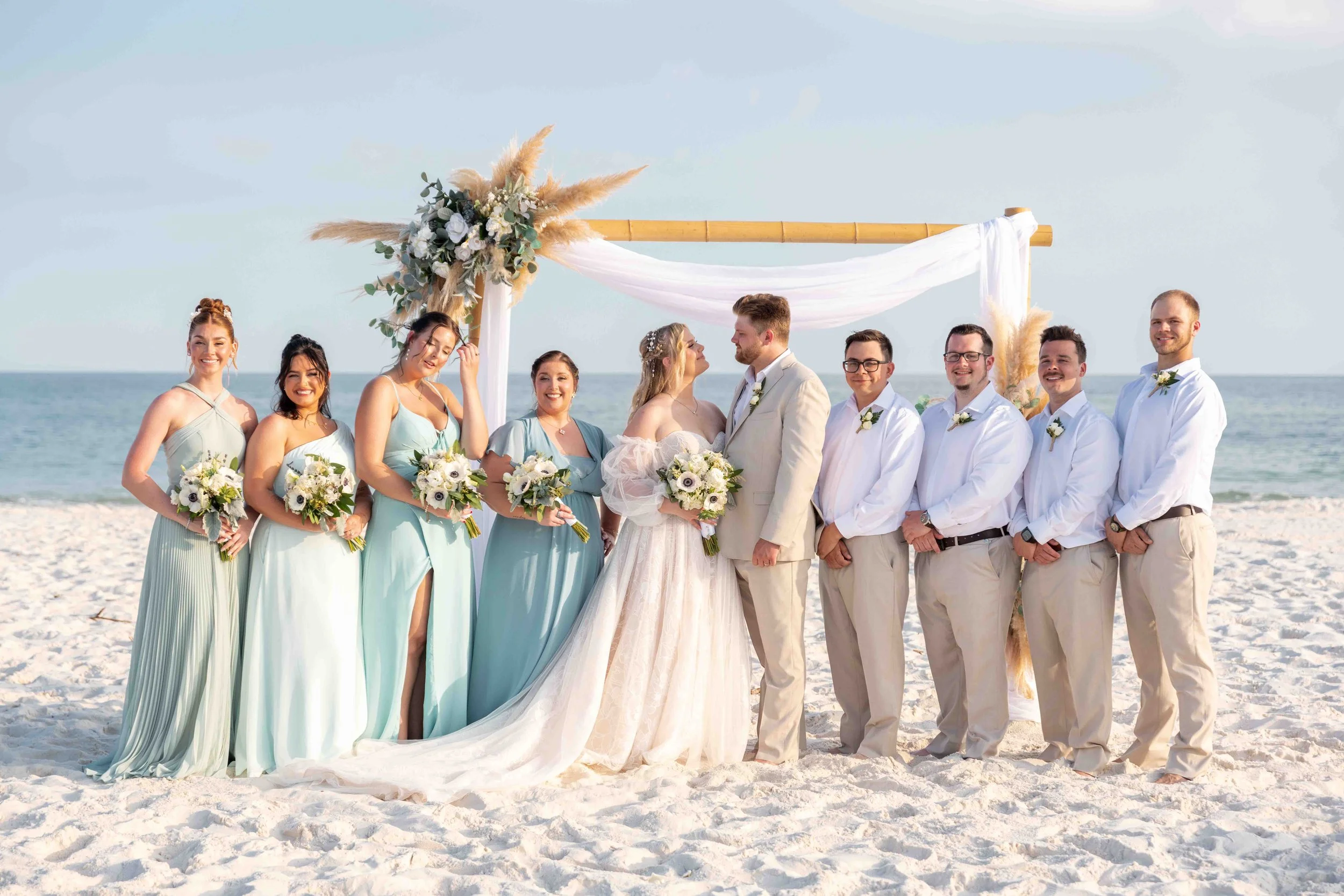 A wedding ceremony on the beach with a bride, groom, bridesmaids, and groomsmen standing under a decorated arch on the sandy shore with the ocean in the background.