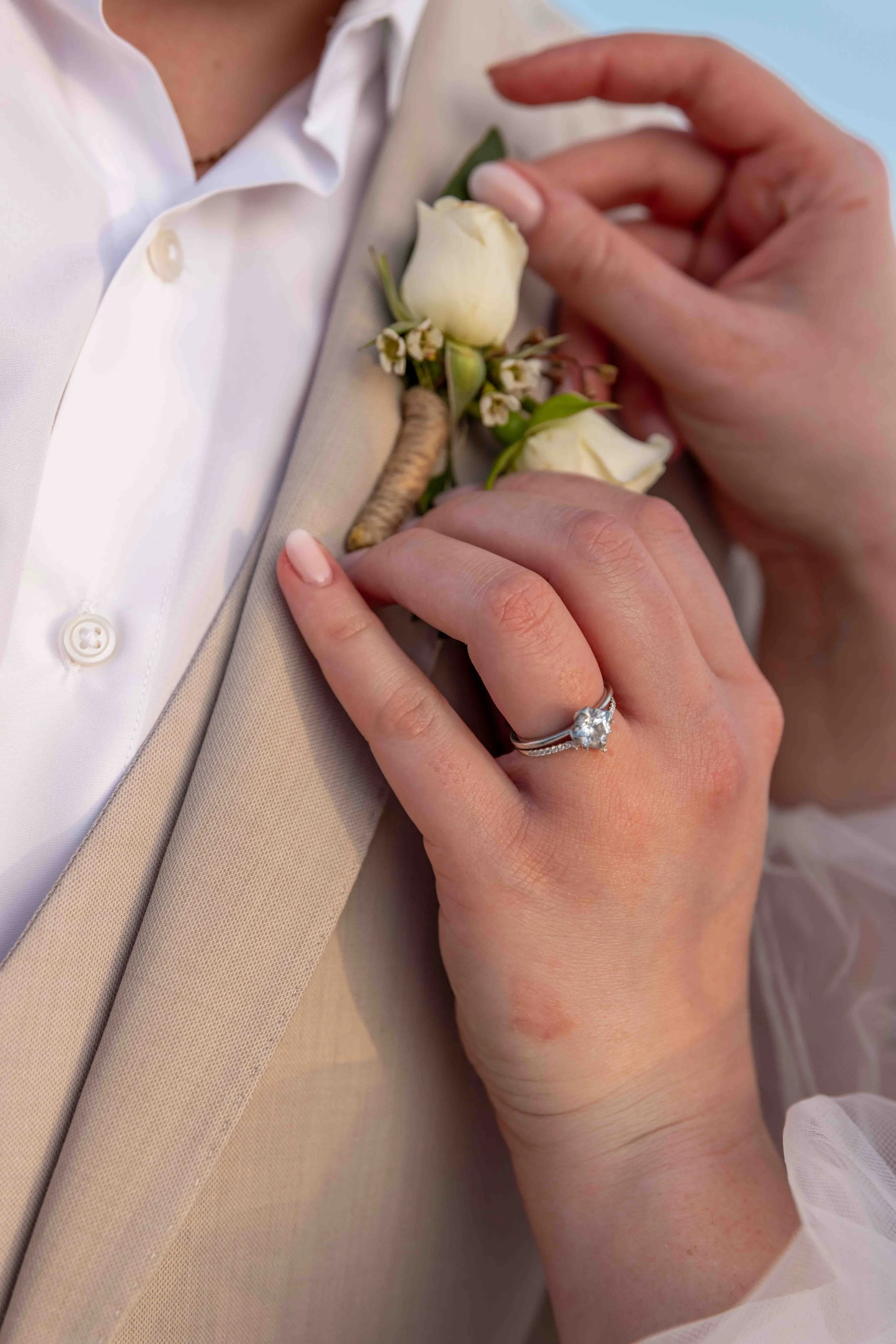 Close-up of a person being pinned with a white rose boutonniere on beige suit jacket, with a wedding ring on their finger.
