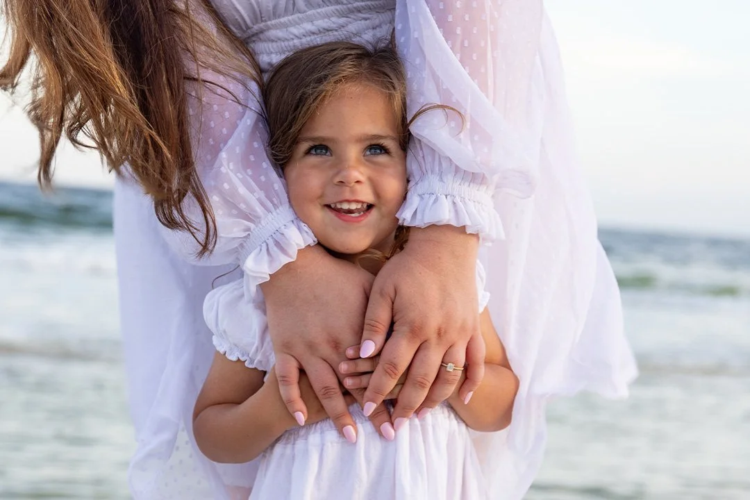 A young girl with blue eyes and a bright smile being embraced by an adult, likely her mother, on a beach with ocean waves in the background.