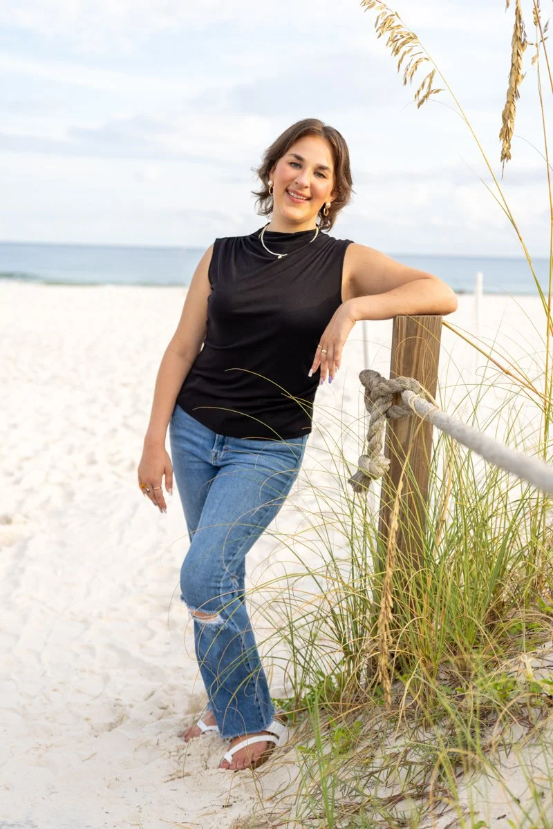 A woman standing at the beach, leaning on a wooden post with a rope, wearing a black sleeveless top, ripped jeans, white sandals, and jewelry, smiling at the camera with the ocean and sky in the background.